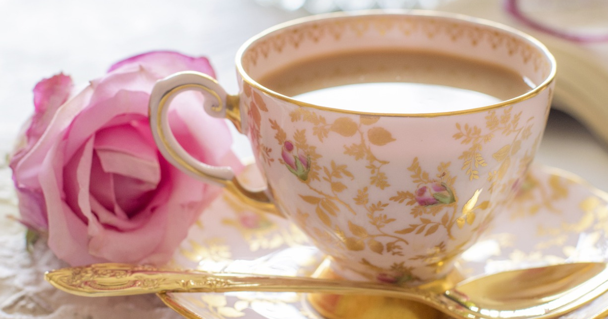 Fine bone china teacup with a gold floral pattern. There is a gold teaspoon on the saucer and a pink rose next to it on the table.